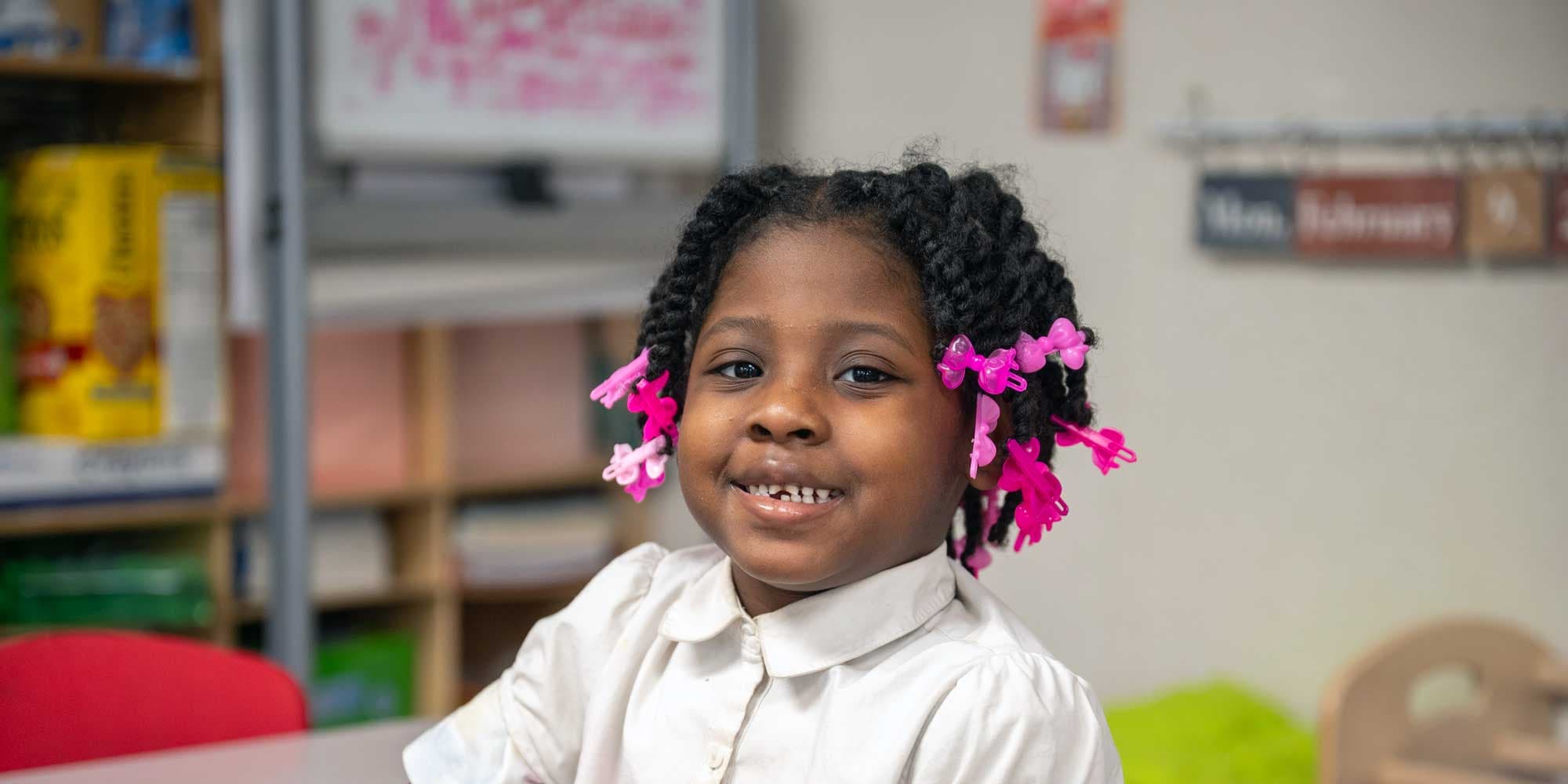 Young student with pink hair clips smiling in a classroom.