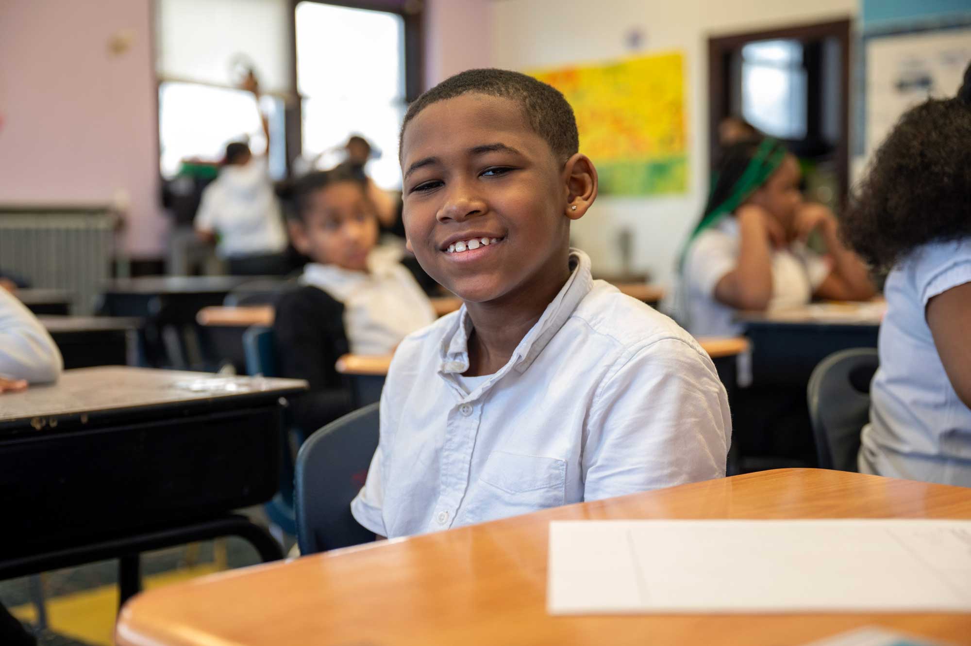 Smiling student seated at a desk in a busy classroom.